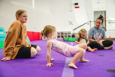 Two young children practicing gymnastics with the guidance of two instructors in a brightly colored gym, focusing on technique and balance.