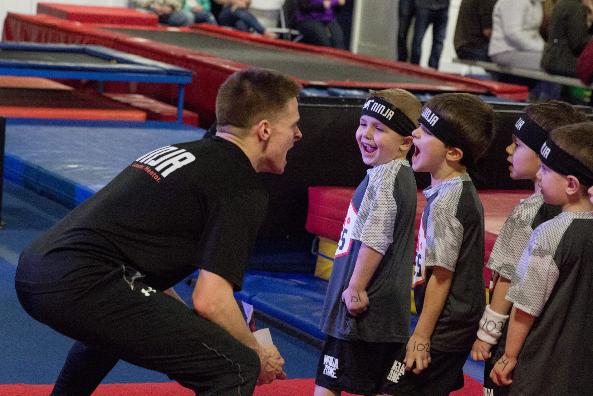 Coach in black Ninja Zone shirt enthusiastically motivating a line of young children wearing Ninja Zone headbands and jerseys at a gymnastics center.
